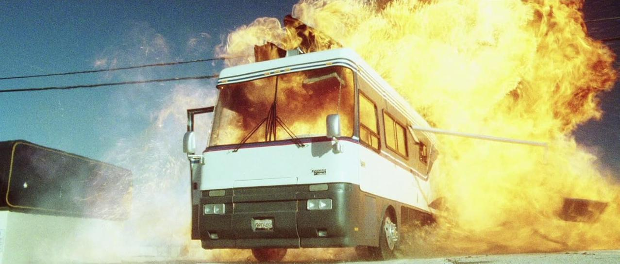 Ein Wohnmobil steht in Flammen. Feuer bricht aus Dach und Seiten, mit blauem Himmel im Hintergrund. Die Front des Fahrzeugs ist sichtbar.