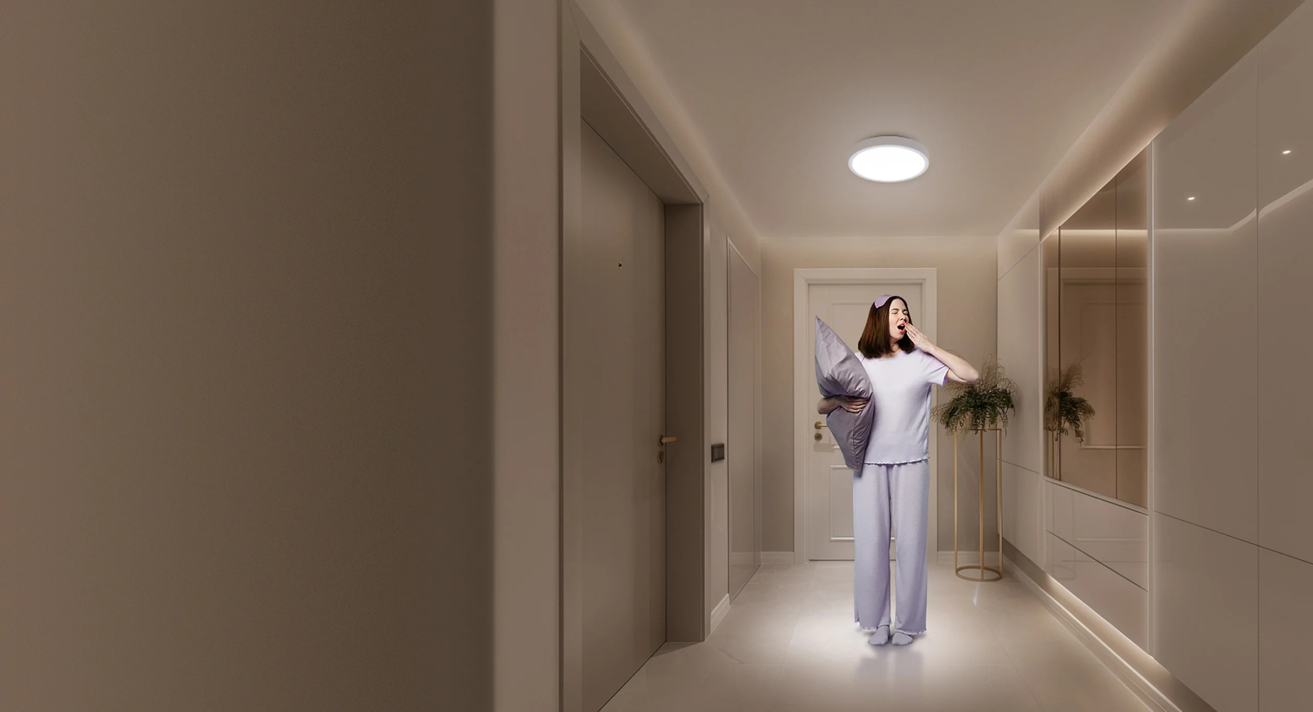 Kobieta ziewa na korytarzu, trzymając poduszkę. Ma na sobie piżamę. Korytarz ma drzwi, światła i roślinę dekoracyjną.
