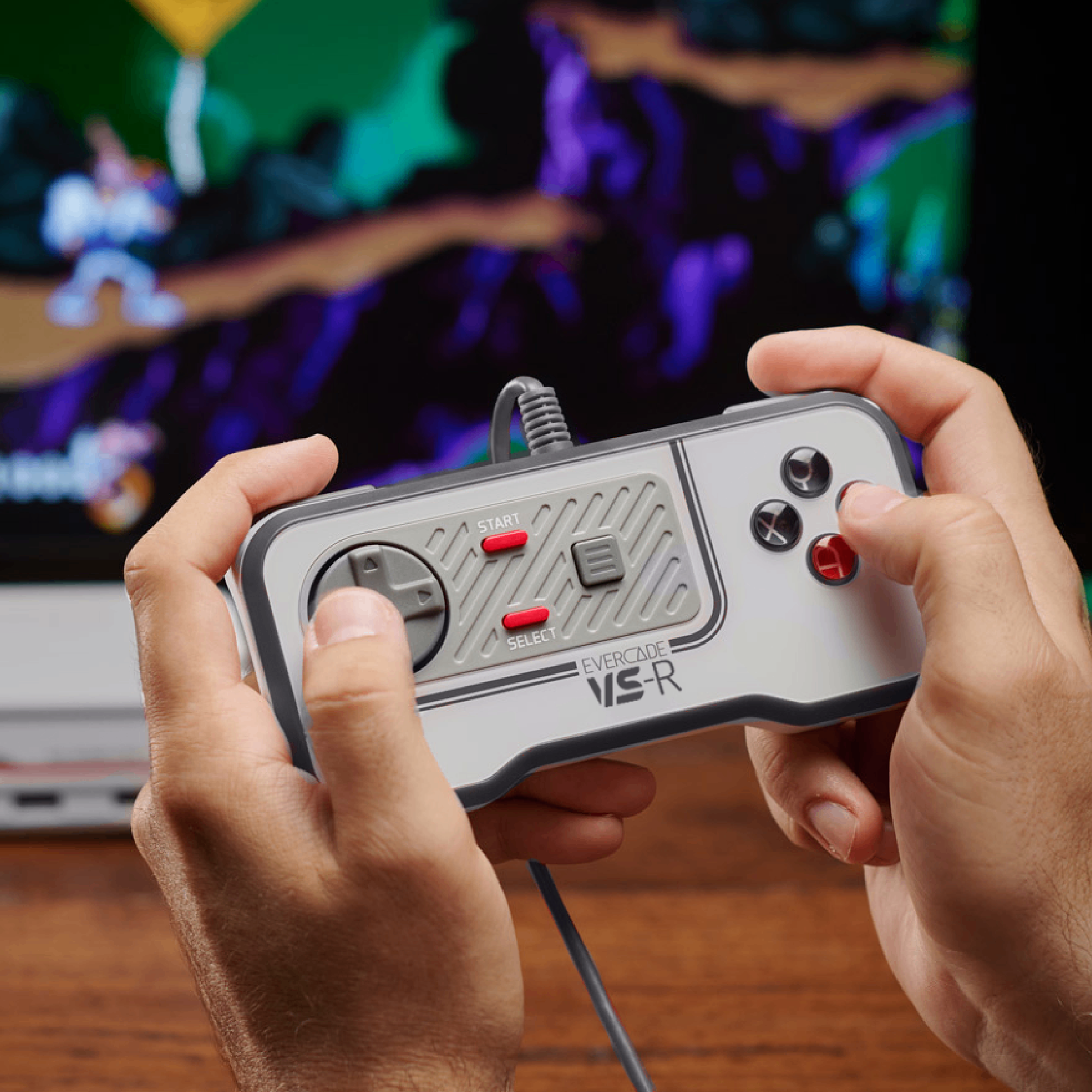 Ręce trzymają kontroler Evercade VS-R. W tle wyświetlana jest gra. Kontroler ma szare i czerwone przyciski.