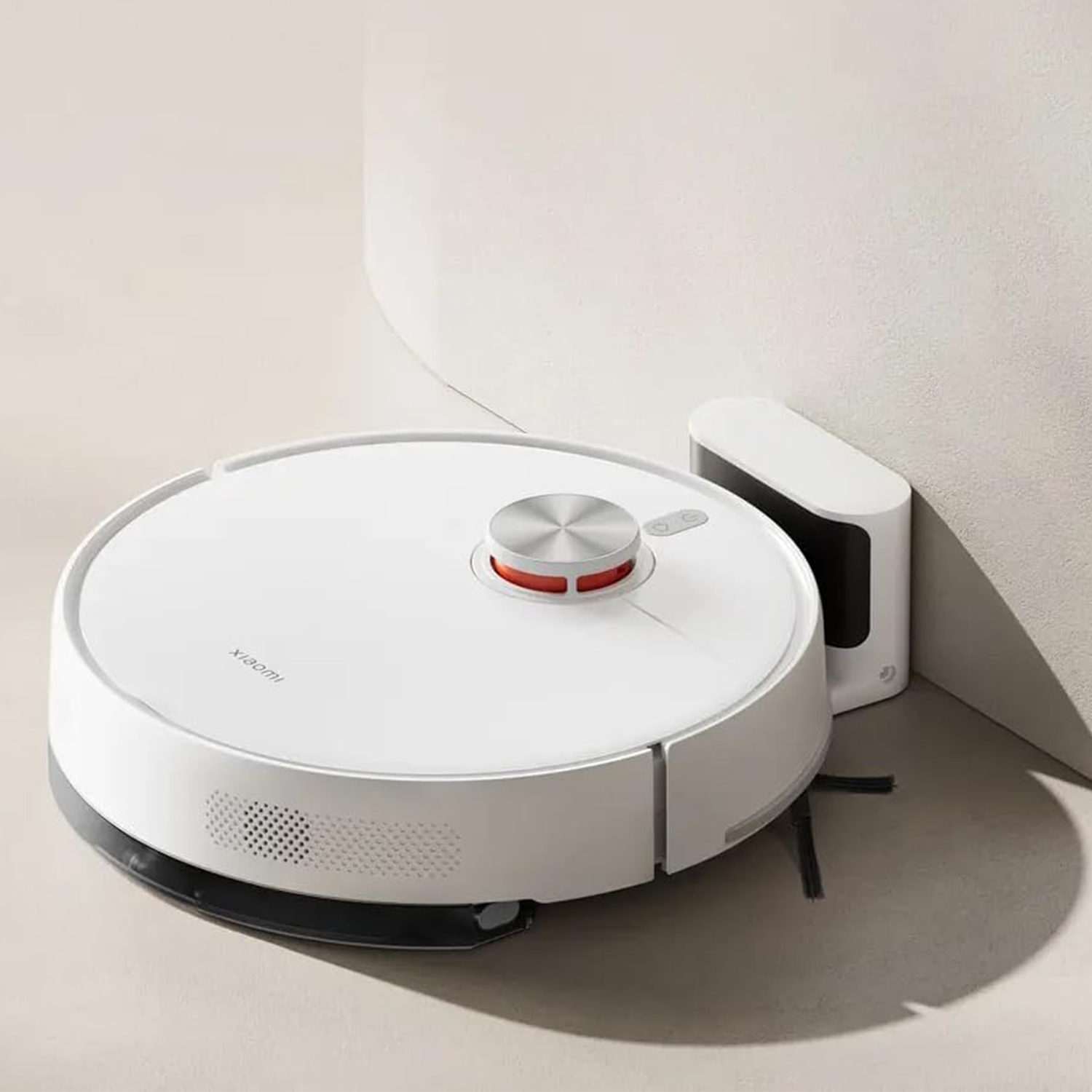 Una aspiradora robot blanca junto a su estación de carga. La aspiradora tiene una parte superior plateada y detalles en rojo. El fondo es beige.