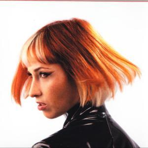 Eine Frau mit orange-blondem Haar, trägt ein schwarzes Oberteil und schaut nach links. Der Hintergrund ist weiß.