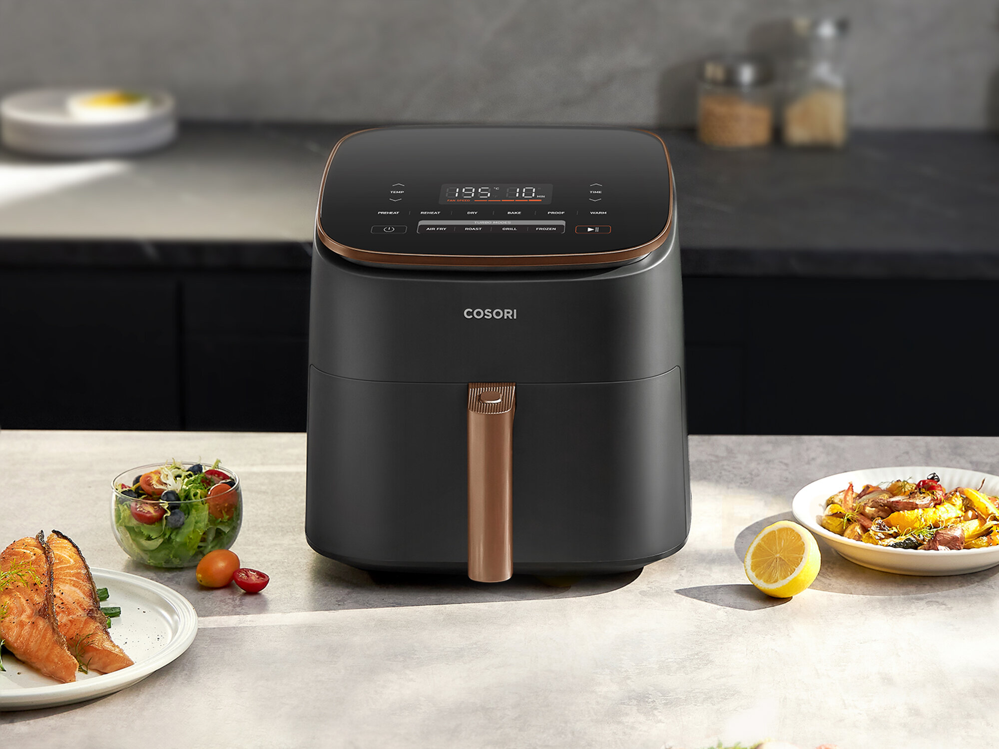 Frytkownica beztłuszczowa stoi na blacie. W pobliżu znajdują się dania z jedzeniem, w tym sałatka i łosoś. Cytryna leży na blacie.