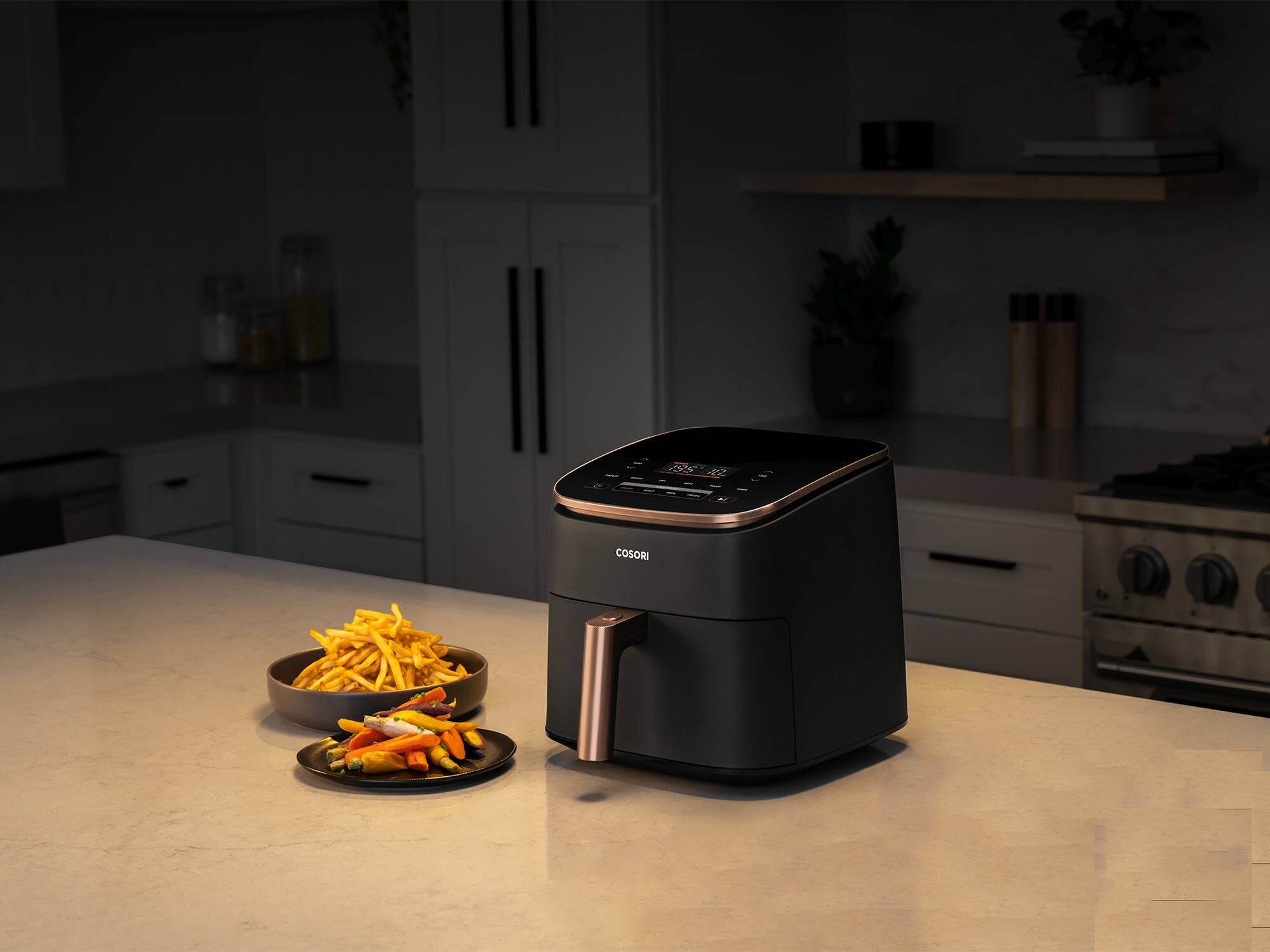 Frytownica beztłuszczowa, frytki i warzywa na blacie kuchennym. Frytownica jest czarna ze złotymi akcentami. Tło obejmuje szafki.