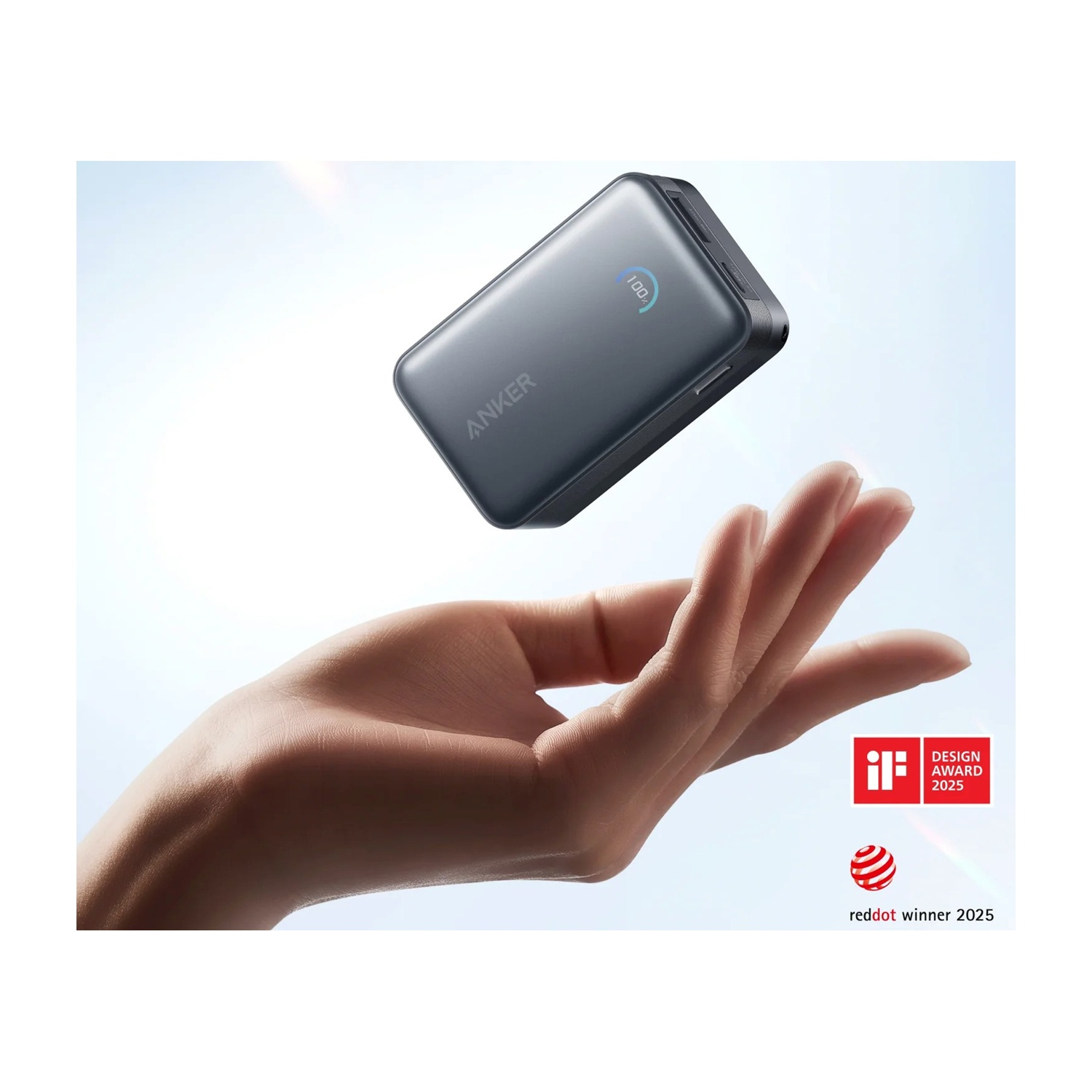Una mano che si protende verso un power bank Anker grigio scuro fluttuante. Mostra '100%'. Premi di design sullo sfondo.
