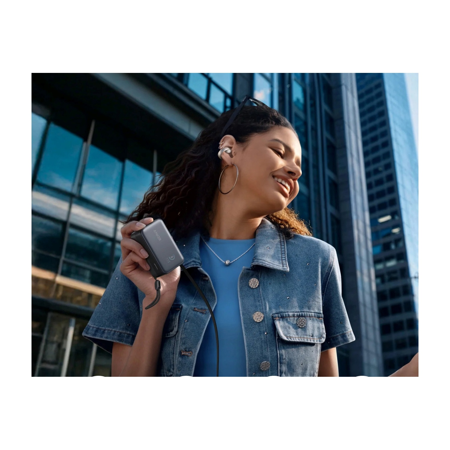 Una donna con auricolari e giacca di jeans sorride, tenendo in mano un piccolo dispositivo grigio. Edifici sullo sfondo riflettono il cielo.