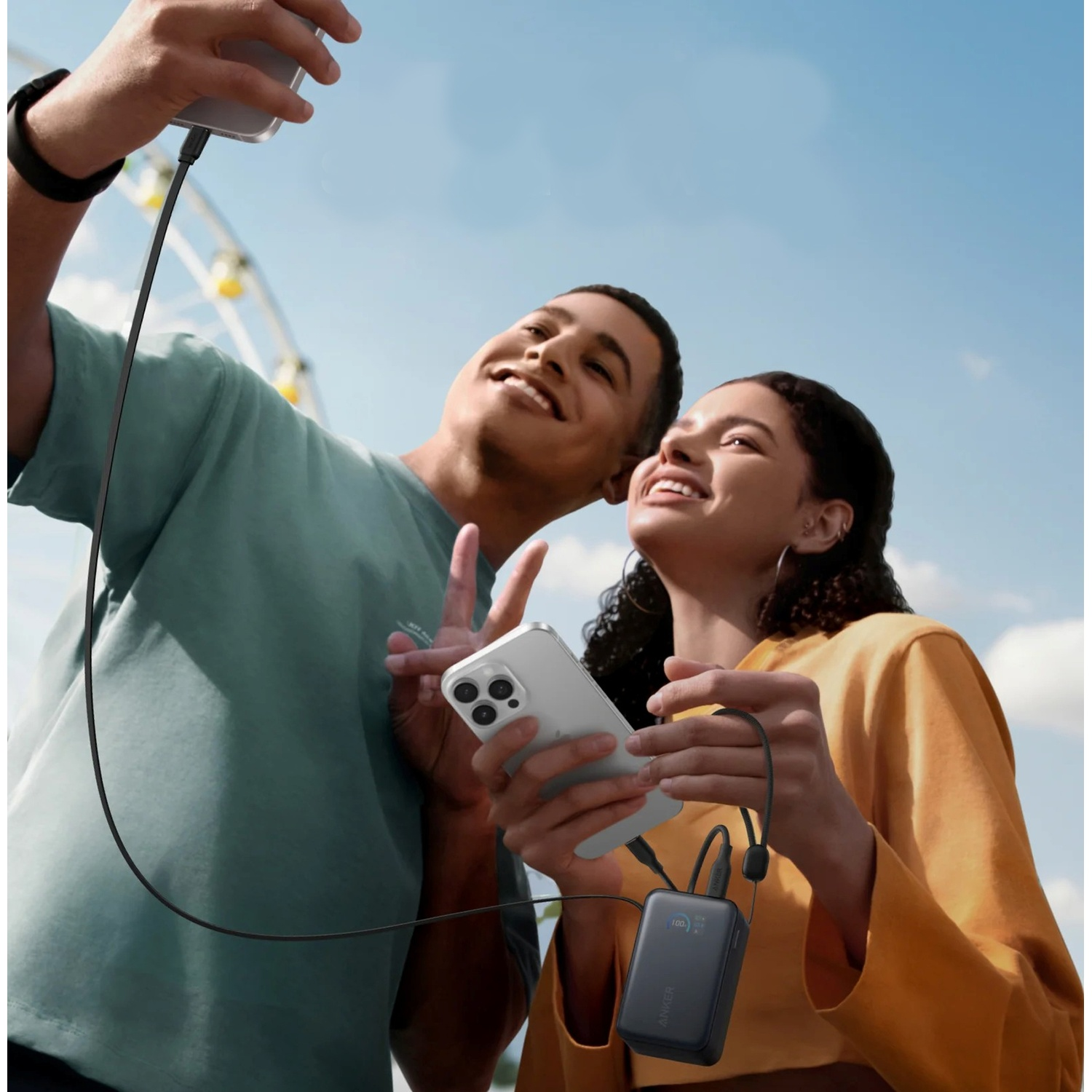 Due persone sorridenti che scattano un selfie con telefoni e un power bank sotto un cielo blu vicino a una ruota panoramica.