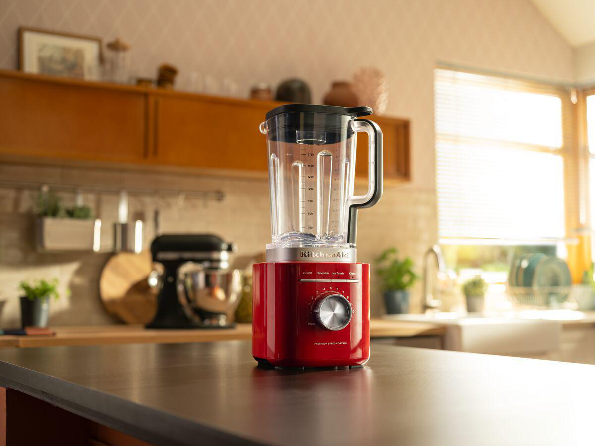 Ein roter KitchenAid-Mixer steht auf einer Küchentheke. Im Hintergrund befindet sich ein schwarzer Mixer. Die Küche hat Holzschränke und ein Fenster.