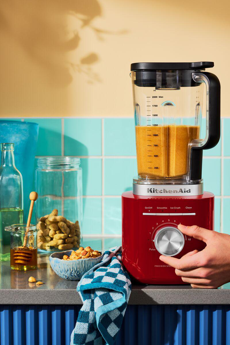 Ein roter Mixer mit orangefarbener Flüssigkeit. Eine Hand dreht den Knopf. Erdnüsse, Honig und ein blaues Handtuch im Vordergrund.