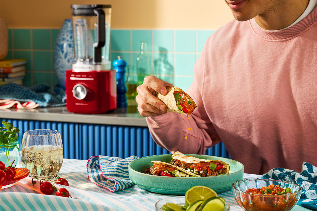 Eine Person hält einen Taco mit Füllung, weitere Tacos in einer Schüssel, Limetten, Salsa und einen roten Mixer im Hintergrund.