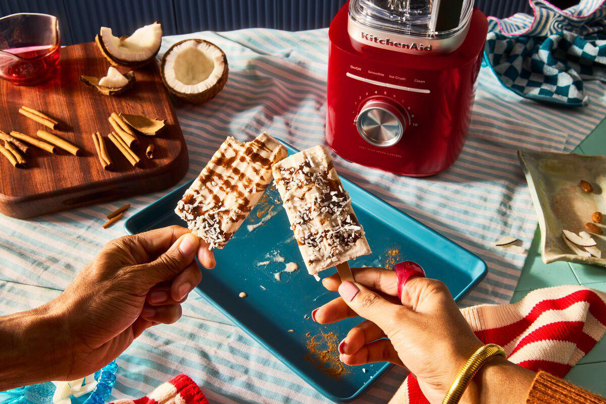 Hände halten zwei Eiscremestiele. Kokosnuss, Zimt, ein roter Mixer und ein rot-weiß gestreiftes Handtuch im Hintergrund.