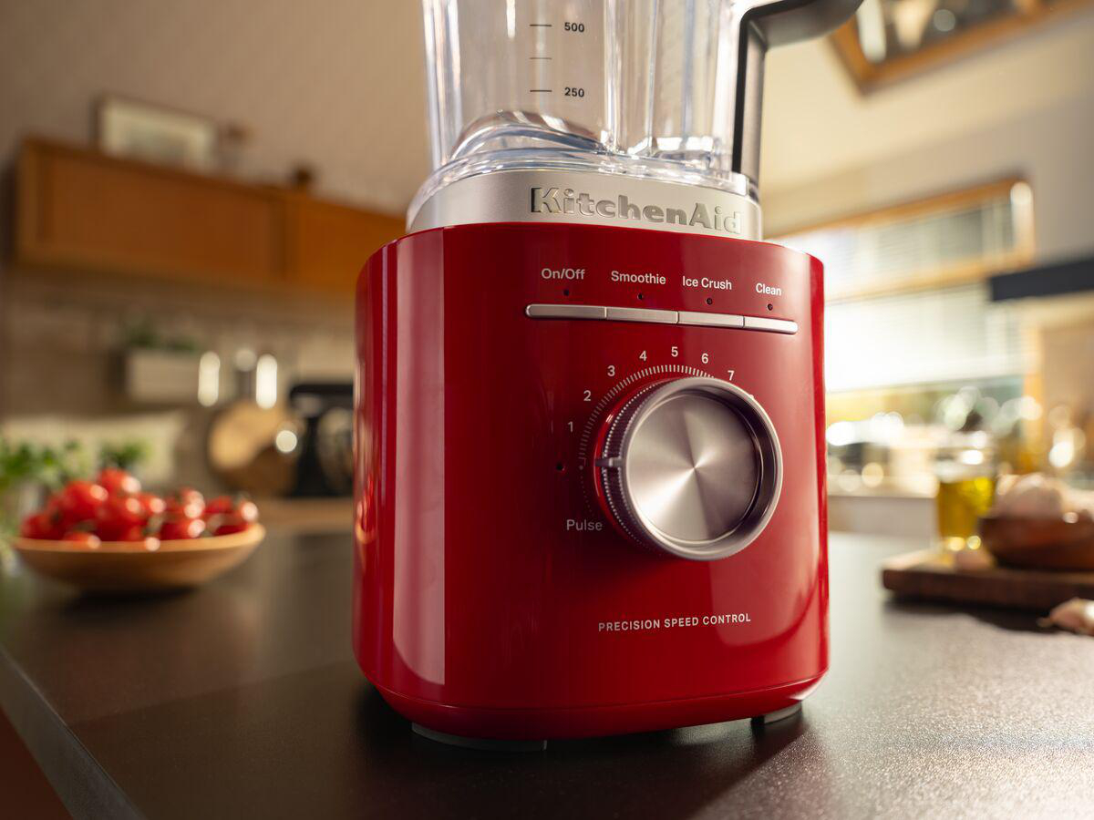 Ein roter KitchenAid-Mixer auf einer Küchentheke. Im Hintergrund befinden sich eine Schüssel mit Tomaten und andere Küchenartikel.