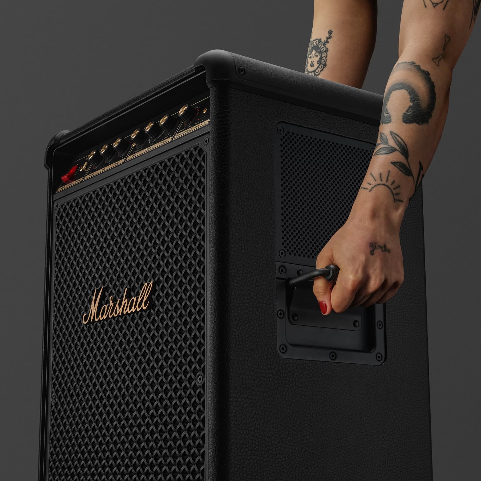 Un amplificateur Marshall noir est tenu par une personne tatouée. L'ampli a une façade en maille avec le logo Marshall.