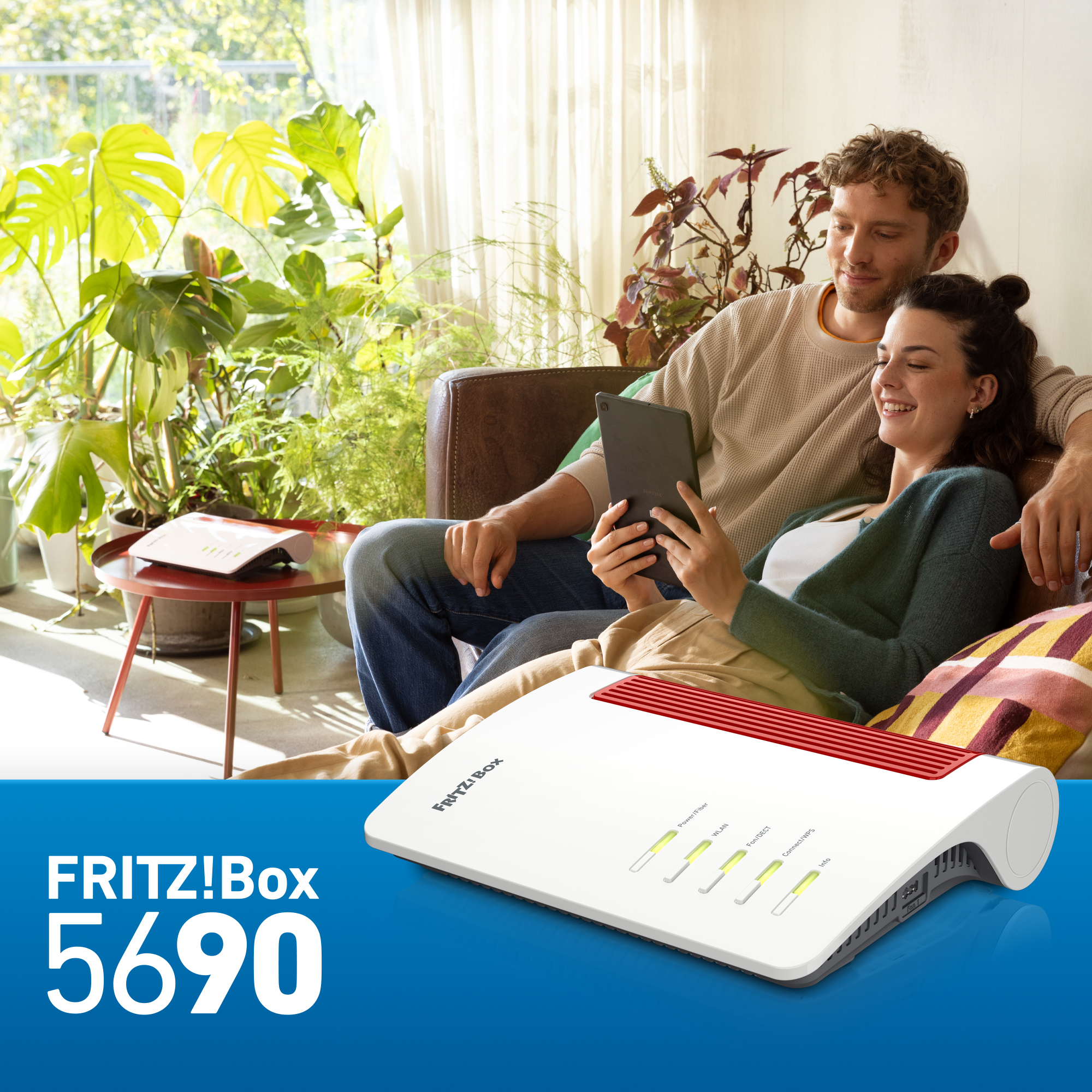 Paar auf einem Sofa, das ein Tablet betrachtet, Router im Vordergrund, umgeben von Pflanzen.