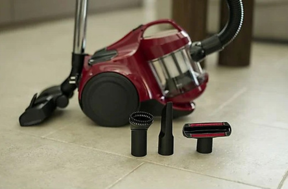 Czerwony odkurzacz z czarnymi nakładkami na jasnej podłodze z płytek. Dodatkowe narzędzia z przodu.