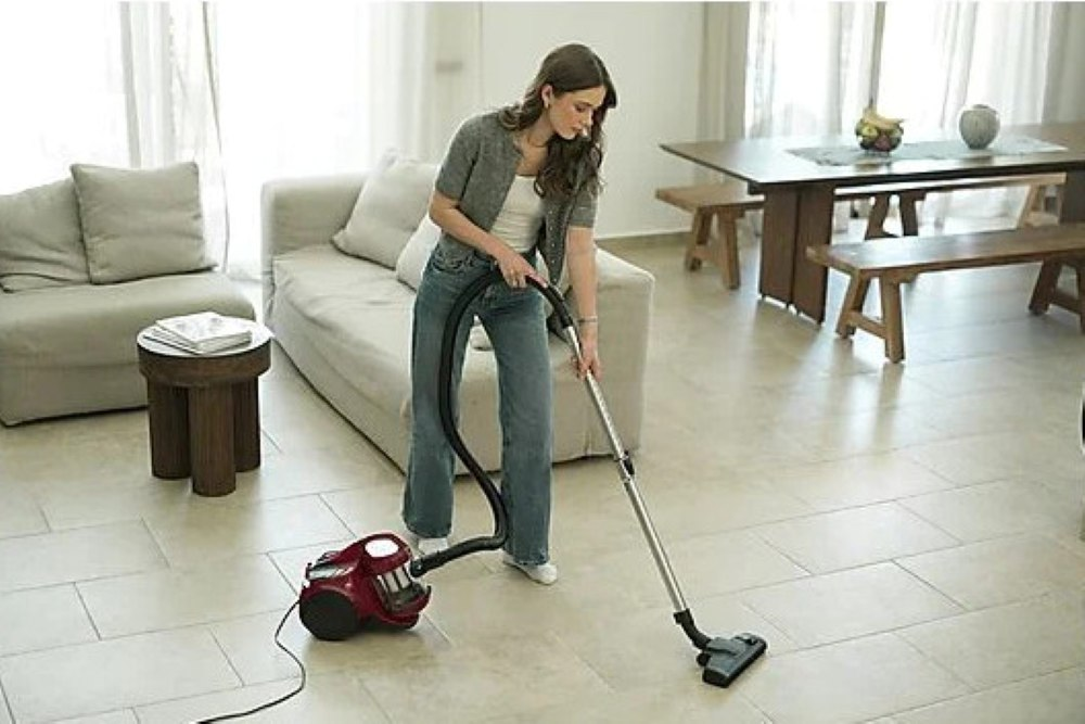 Kobieta odkurza salon. Czerwony odkurzacz stoi na podłodze. W tle kanapa i stół jadalniany.