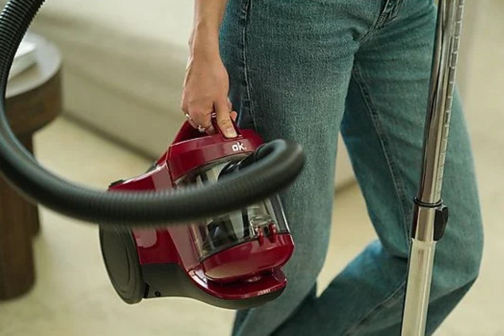 Osoba trzyma czerwony odkurzacz. Widoczne są również dżinsy, czarny wąż i srebrna różdżka, z rozmytym tłem.