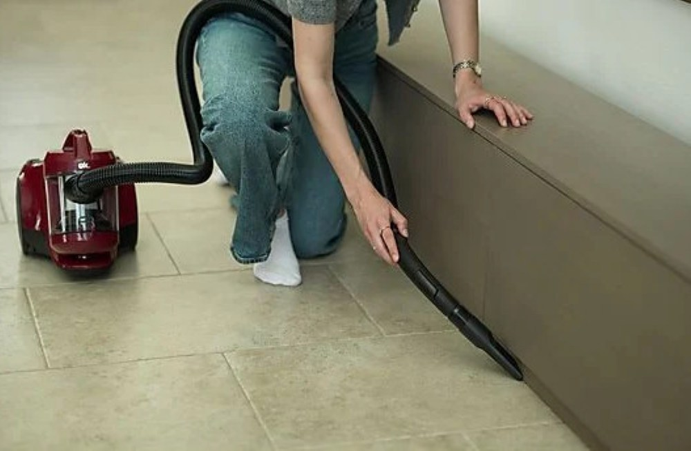 Osoba klęczy, odkurzając podłogę. Czerwony odkurzacz jest podłączony do węża. Odkurzają podstawę brązowej szafki.