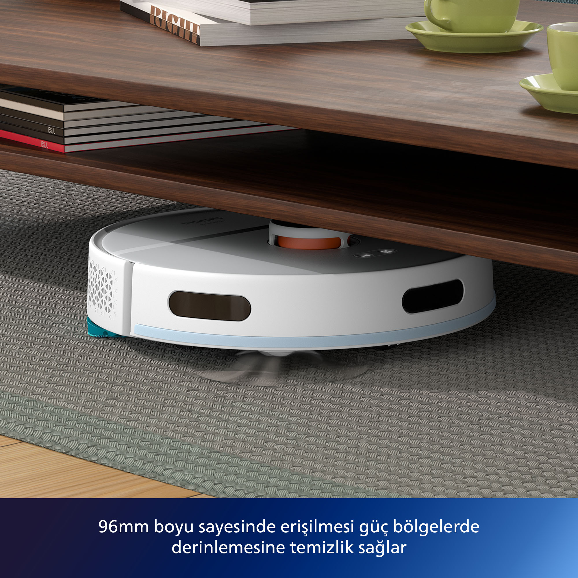 Kahverengi bir masanın altında beyaz ve gri bir robot süpürge. Masanın üzerinde kitaplar ve yeşil fincanlar var.