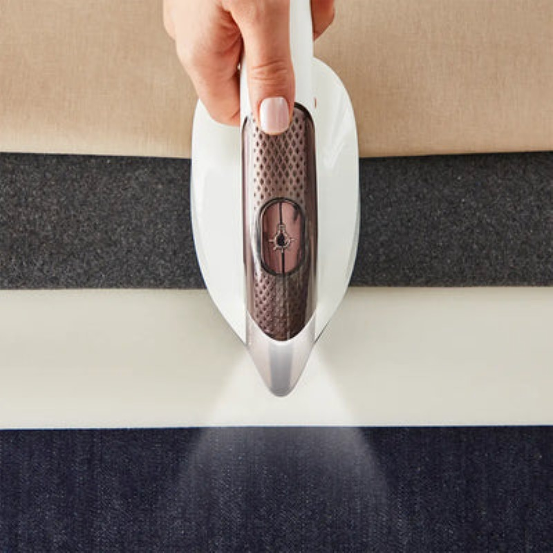 Dłoń trzyma białe żelazko nad tkaniną. Para wydobywa się z żelazka. Tkaniny są beżowe, szare, białe i granatowe.