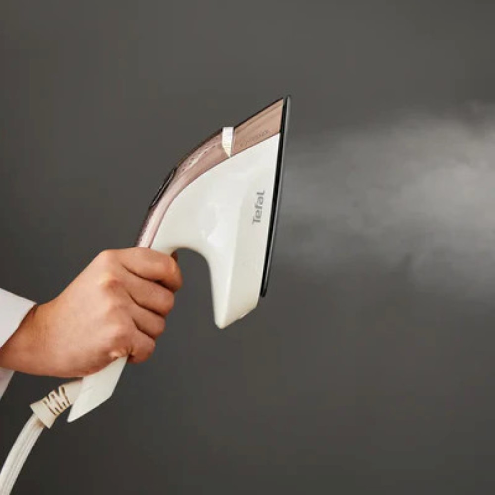 Dłoń trzyma biało-złote żelazko, wydobywa się para. Tło jest gładkie szare.