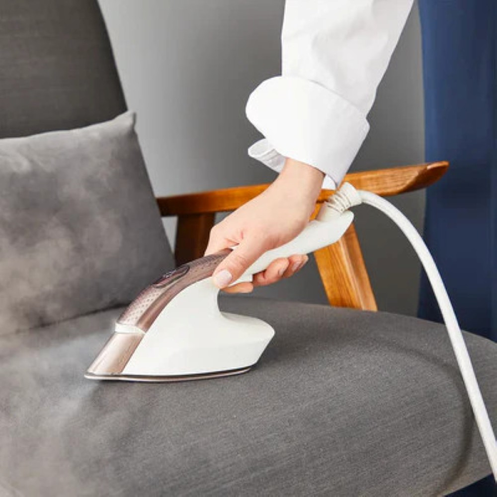 Osoba prasuje szary fotel biało-brązowym żelazkiem parowym. Para unosi się z żelazka. Osoba ma na sobie białą koszulę.