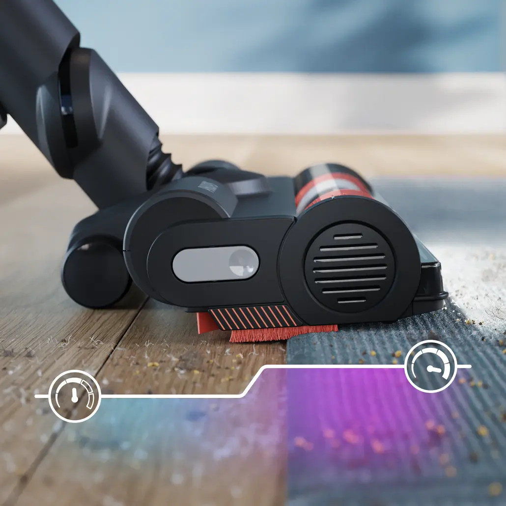 Głowica odkurzacza na drewnie i dywanie. Ma czarny i szary korpus z czerwonym włosiem. Dywan jest w połowie czysty.