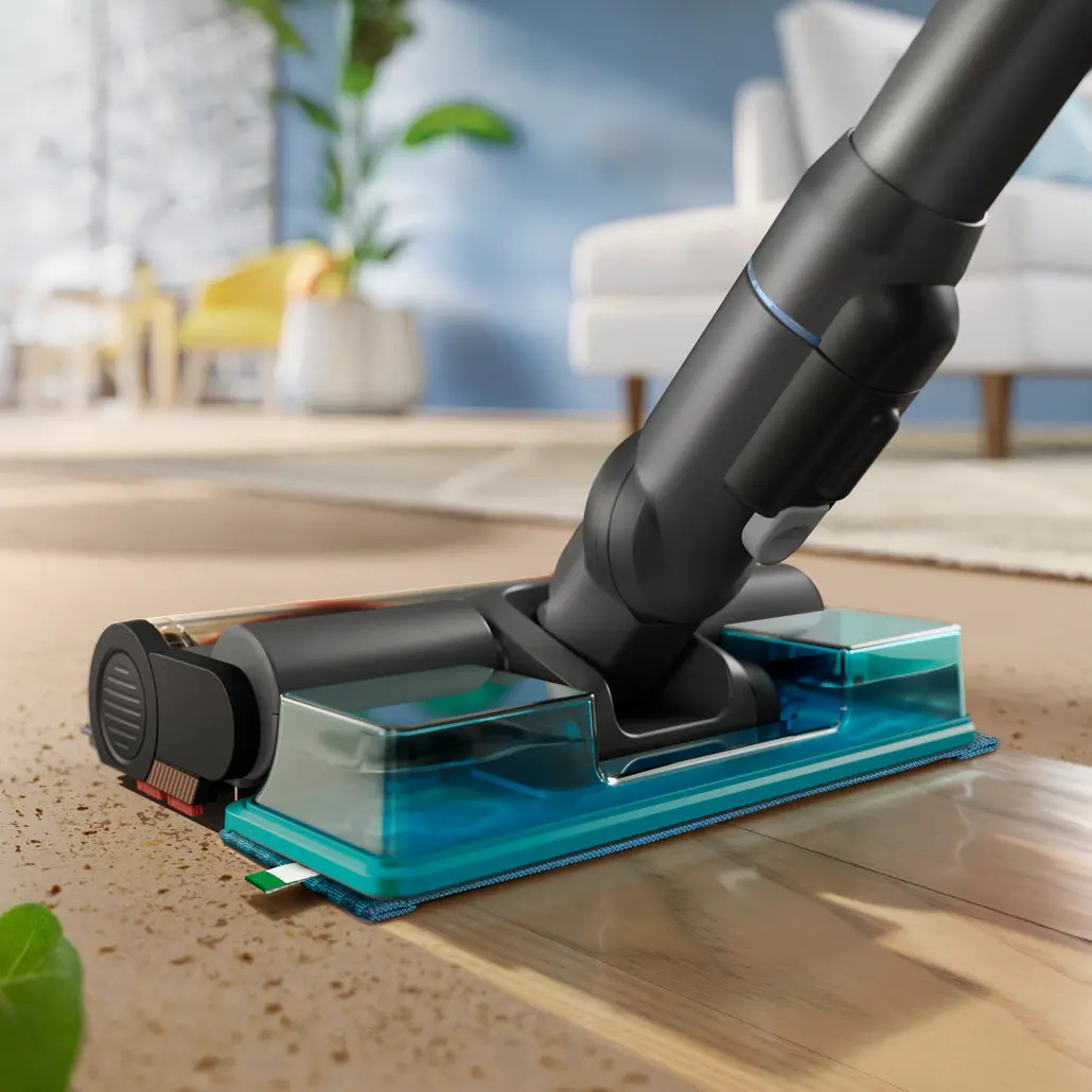 Zbliżenie czarnej i turkusowej głowicy odkurzacza czyszczącej drewnianą podłogę. Kanapa i rośliny są na rozmytym tle.