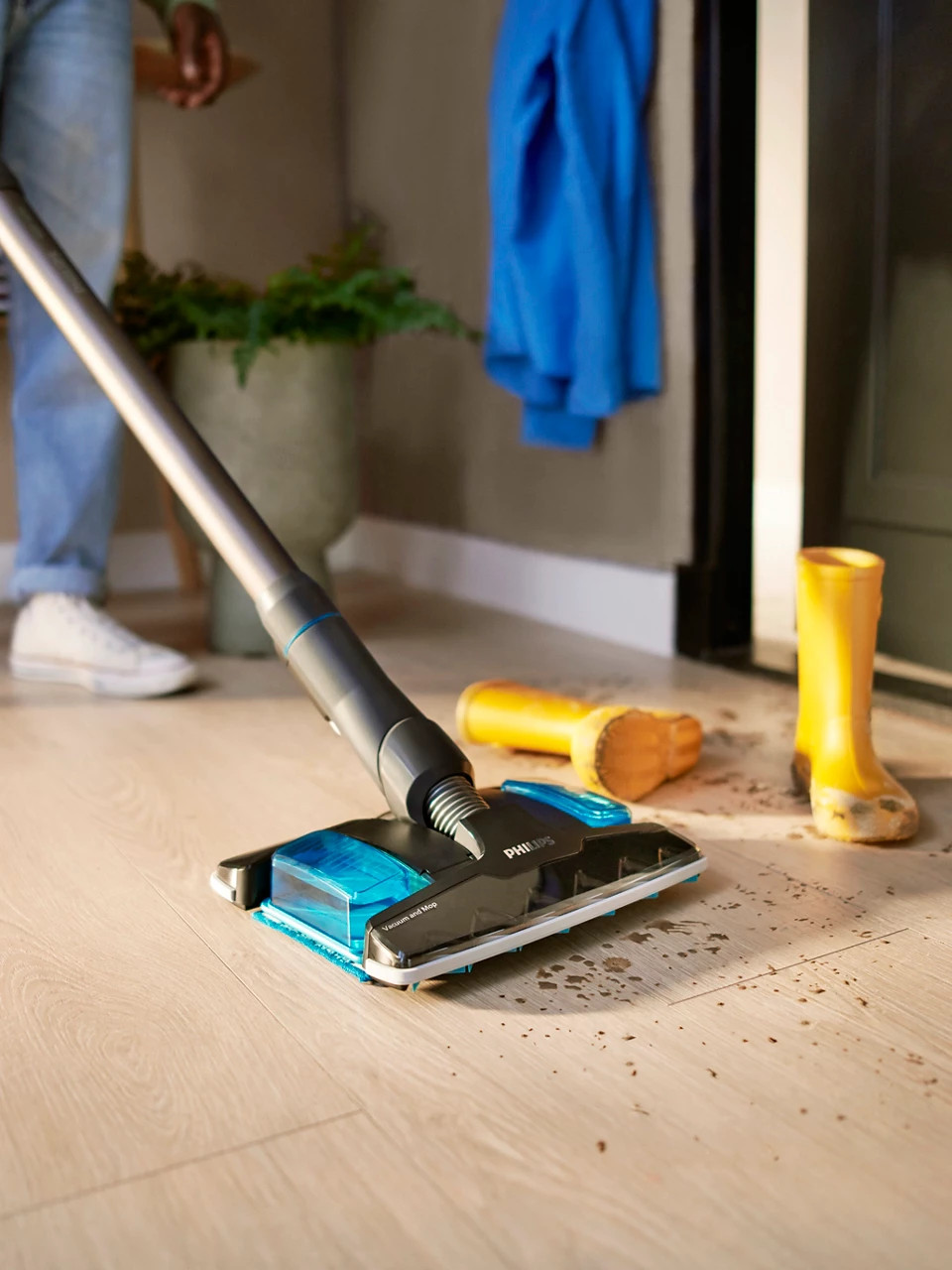 Osoba używająca odkurzacza Philips. Żółte buty, niebieska kurtka i doniczka w tle. Podłoga jest brudna.