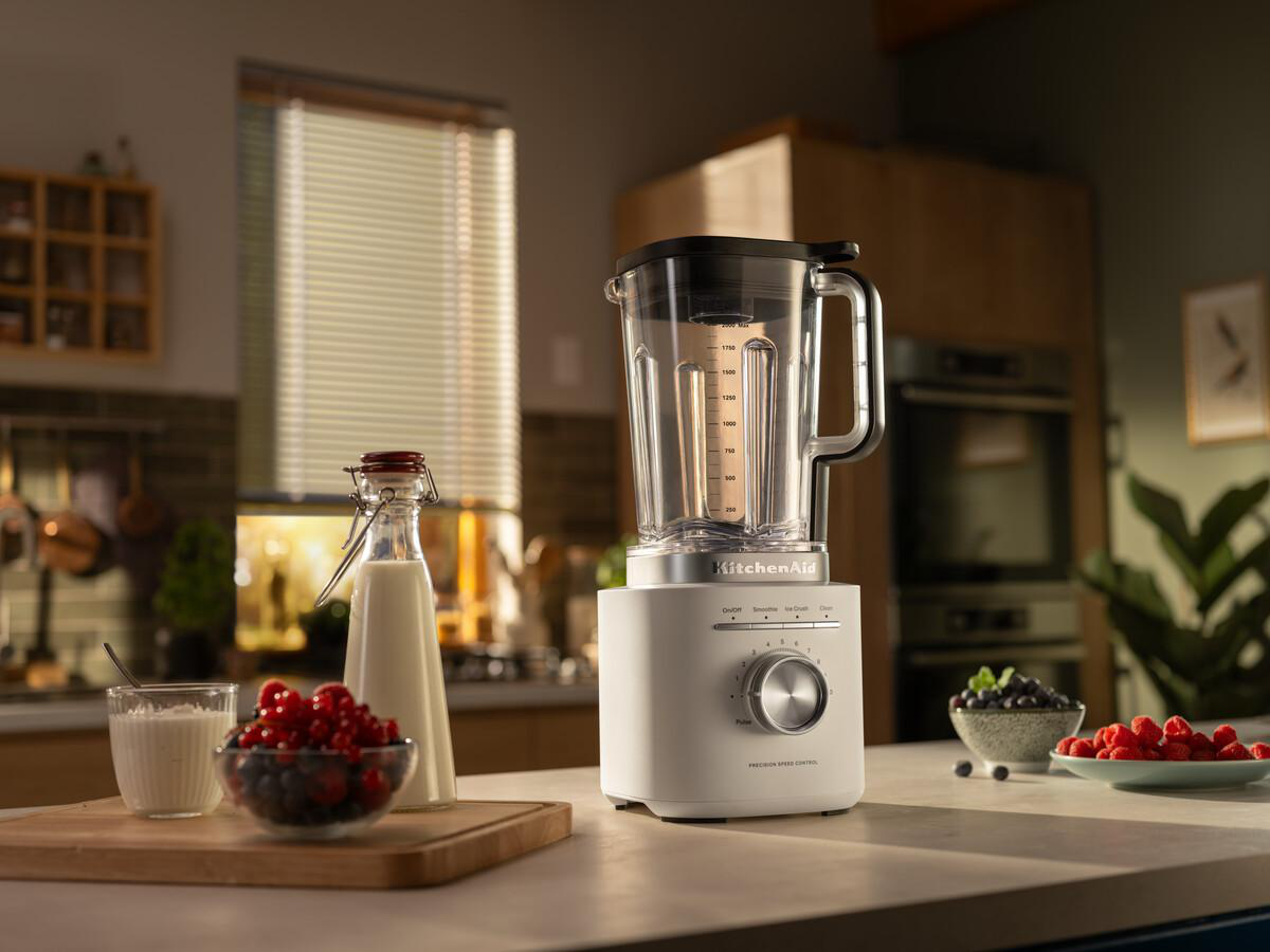 Ein weißer KitchenAid-Mixer auf einer Theke. Milch, Beeren und ein Glas sind im Vordergrund. Ein Fenster und eine Küche sind im Hintergrund.