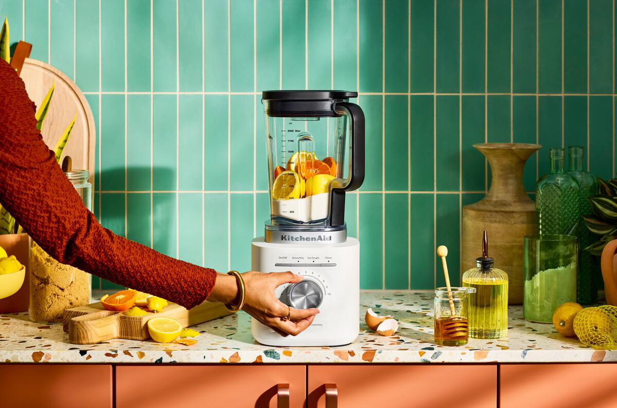 Eine Person benutzt einen KitchenAid-Mixer, gefüllt mit Früchten. Die Szene beinhaltet Honig, Zitronen und einen grünen Fliesen-Hintergrund.