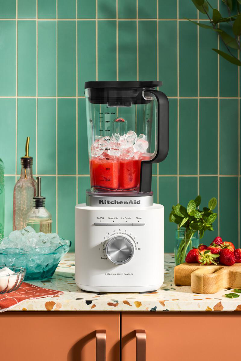 Ein weißer KitchenAid-Mixer mit Eis und roter Flüssigkeit. Erdbeeren und Minze liegen in der Nähe. Türkisfarbene Fliesen im Hintergrund.