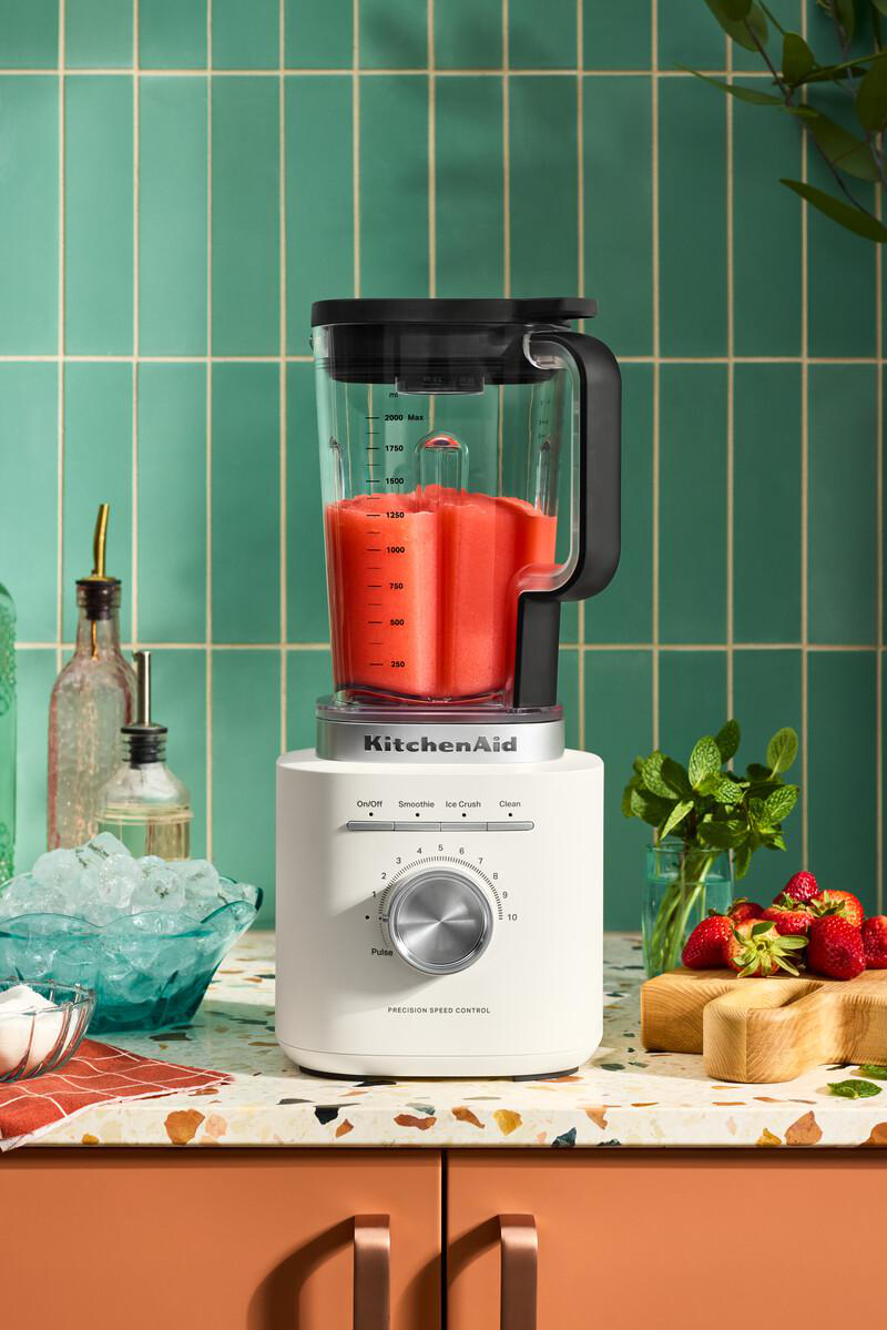 Ein weißer KitchenAid-Mixer mit roter Flüssigkeit, Erdbeeren, Minze und Eis auf einer Arbeitsplatte. Grüner Fliesen-Hintergrund.