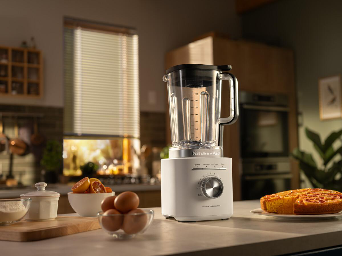 Ein KitchenAid-Mixer steht auf einer Theke mit Zutaten: Orangen, Eiern, Mehl und einem Kuchen. Im Hintergrund ein Fenster mit Jalousien.