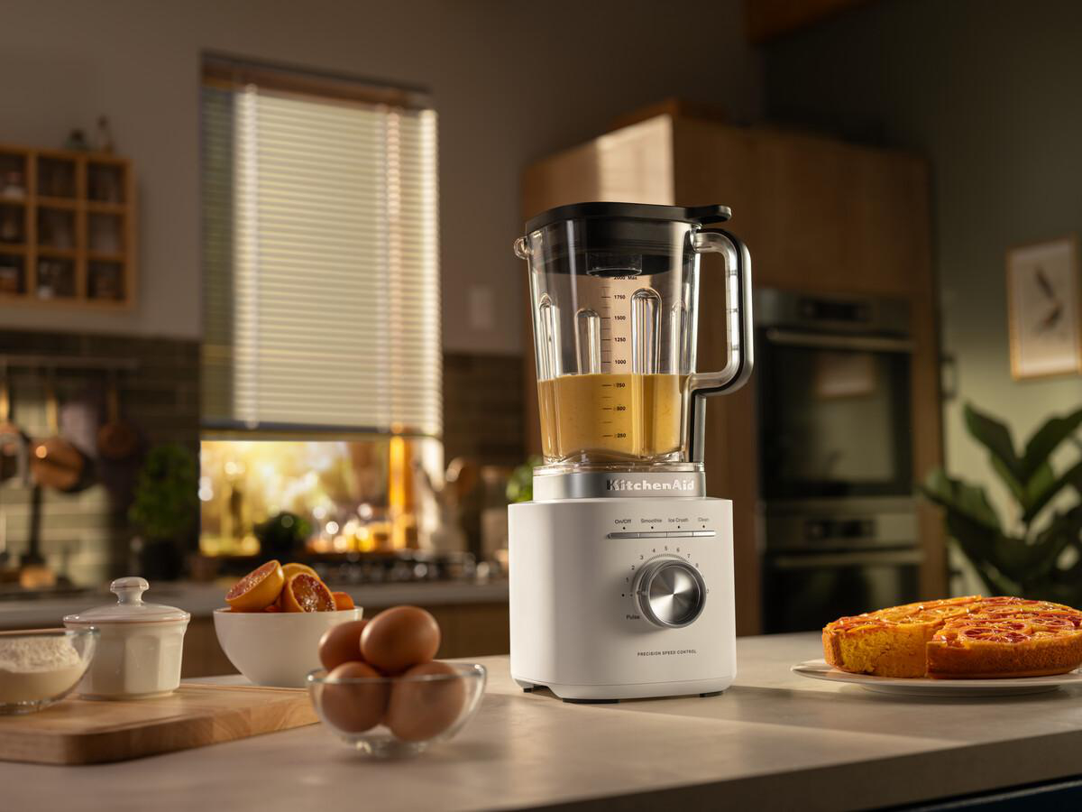 Küchenszene mit Mixer, Kuchen, Eiern und Zitrusfrüchten. Der Mixer steht auf der Arbeitsplatte. Der Hintergrund umfasst ein Fenster und einen Ofen.