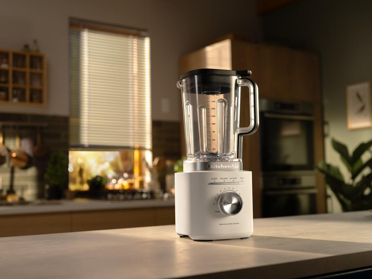 Ein weißer KitchenAid-Mixer steht auf einer Theke. Dahinter eine Küche mit Fenster und Schränken. Die Szene ist gut beleuchtet.