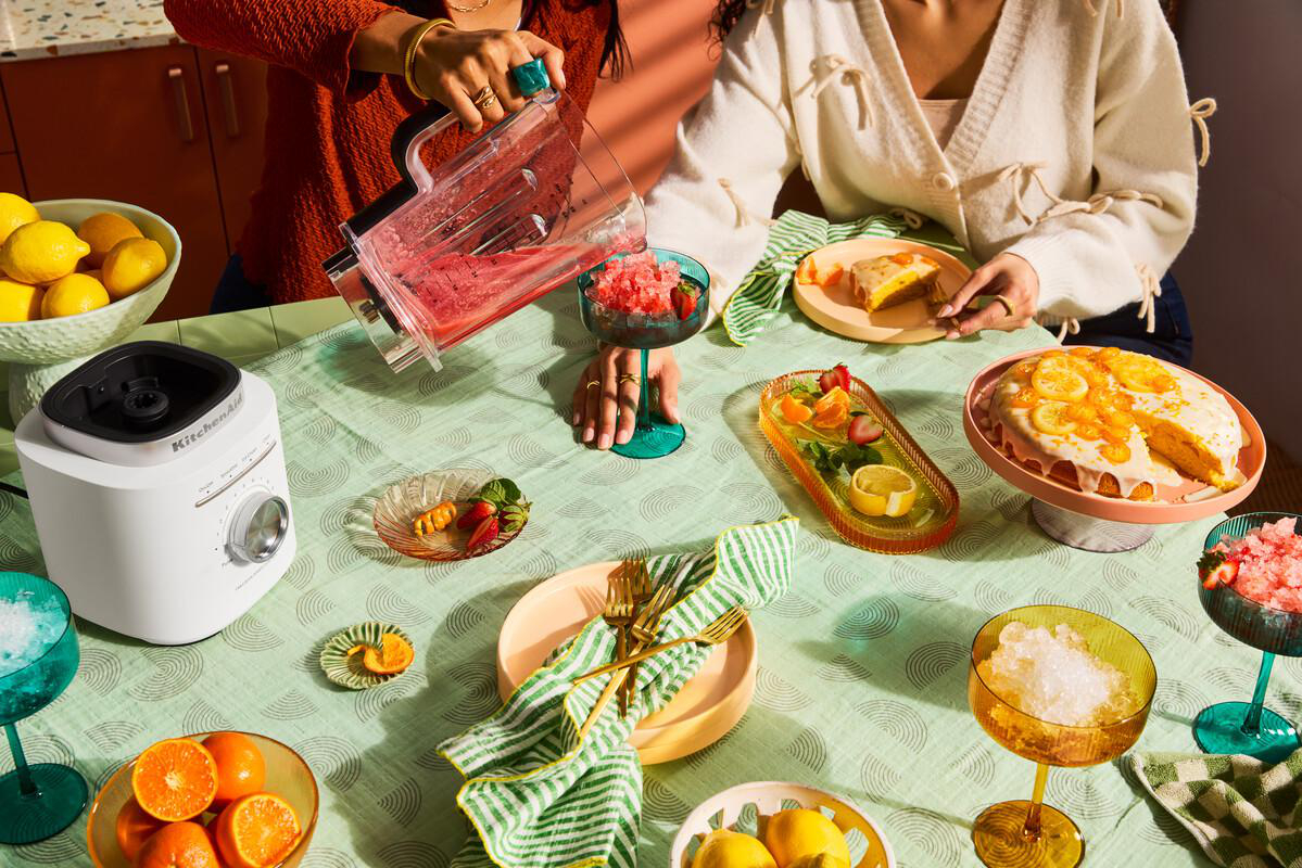 Personen gießen ein gemixtes Getränk in Gläser. Tisch mit Kuchen, Obst und Getränken. Weißer Mixer links.