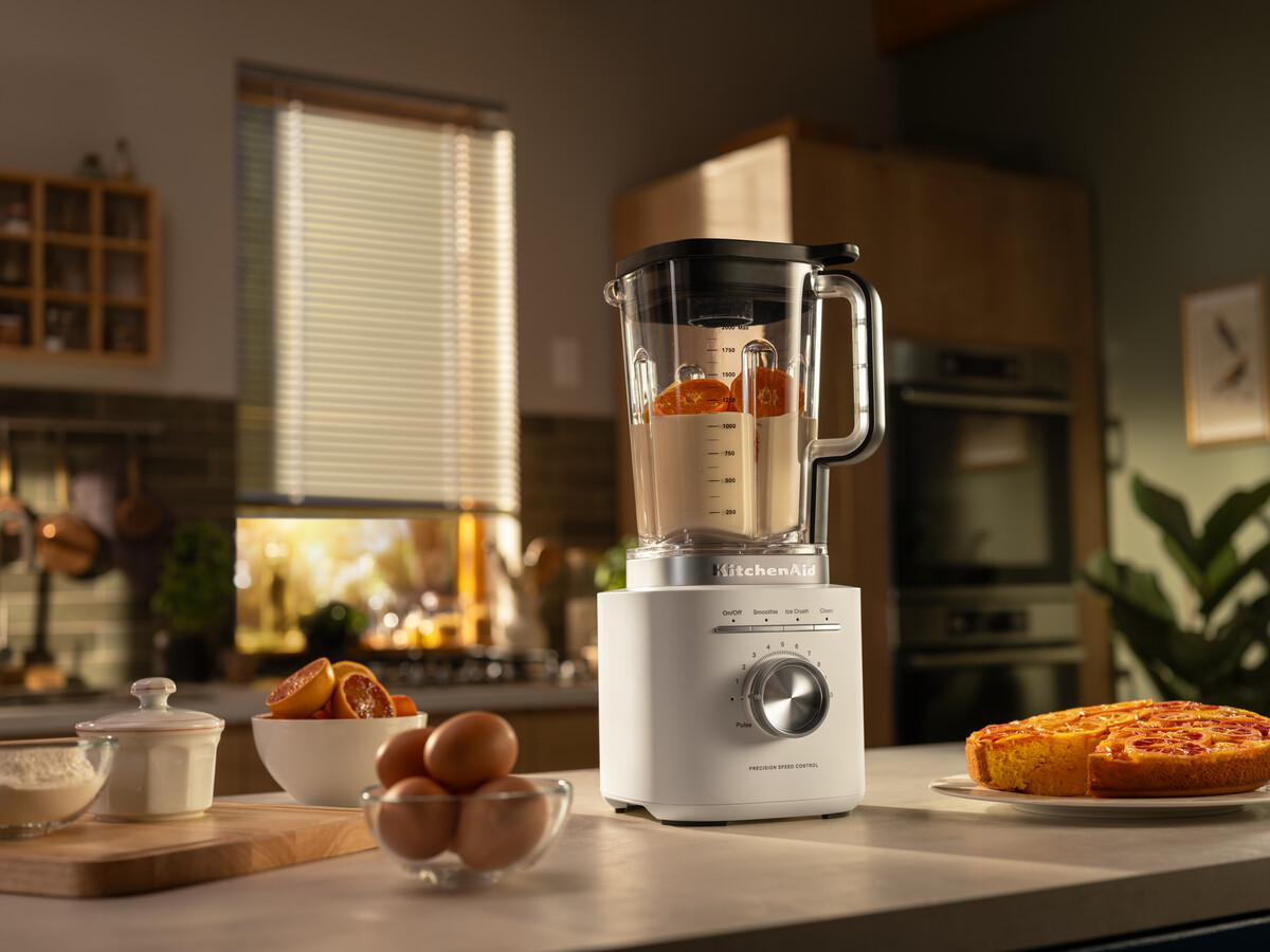 Ein weißer KitchenAid-Mixer mit Orangen. Auf der Theke stehen Schalen mit Eiern, Orangen und einem Kuchen.