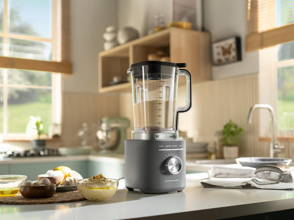Ein KitchenAid-Mixer auf einer Arbeitsplatte. Hummus und Zutaten sind davor. Im Hintergrund ein Fenster und ein Holzregal.