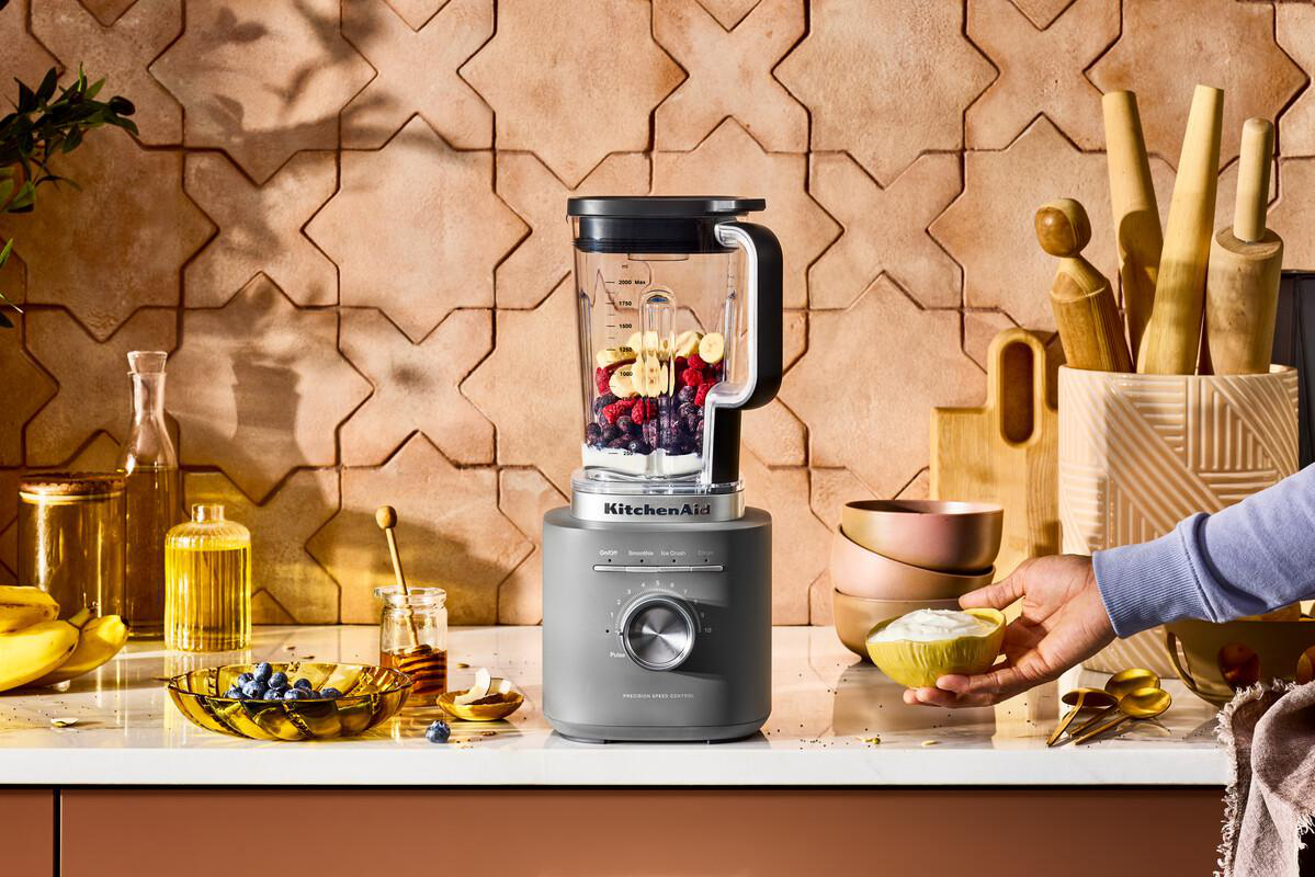 Ein Mixer mit Früchten, eine Person, die Joghurt hält, andere Zutaten und Küchenutensilien auf einer Theke.