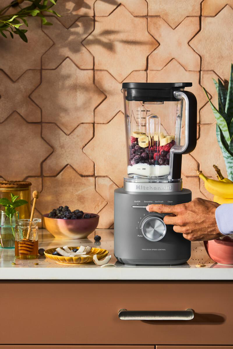Ein Mixer mit Zutaten, eine Hand dreht den Drehknopf. Weitere Früchte und Honig stehen auf der Theke.