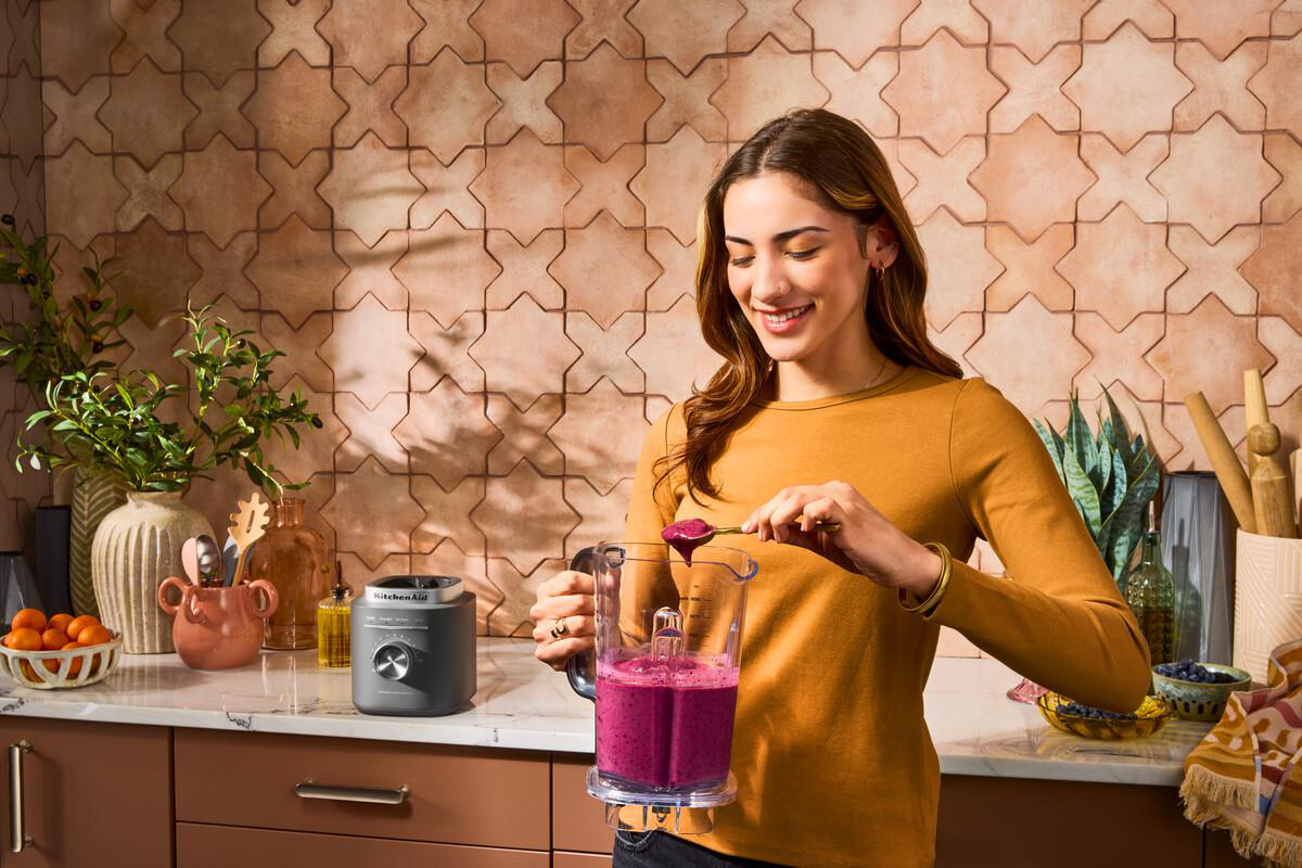 Eine lächelnde Frau mit einem Löffel schöpft Smoothie aus einem Mixer. Küche mit Früchten, Pflanzen und einer gefliesten Wand.