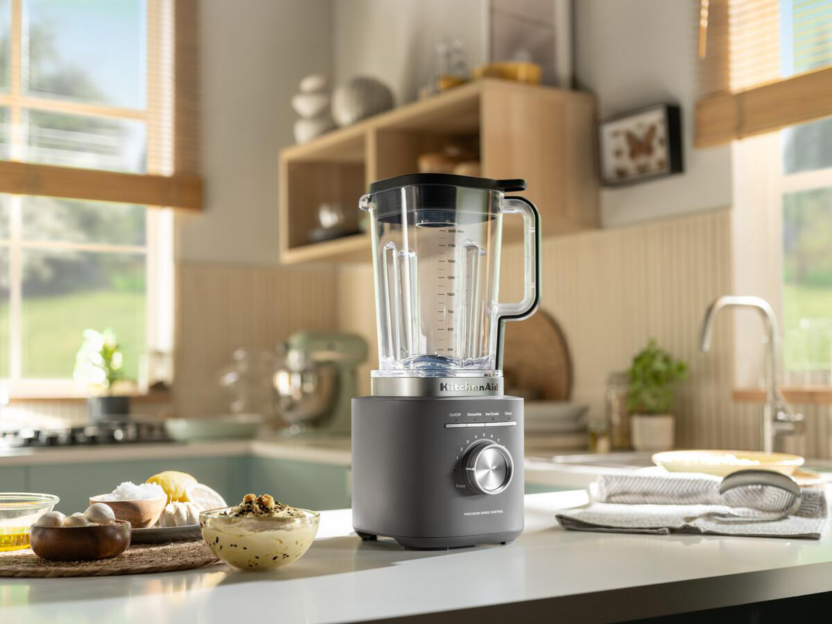 Ein KitchenAid-Mixer steht auf einer Theke. Im Hintergrund sind Schüsseln mit Essen und ein Küchenbecken. Ein Fenster ist ebenfalls sichtbar.