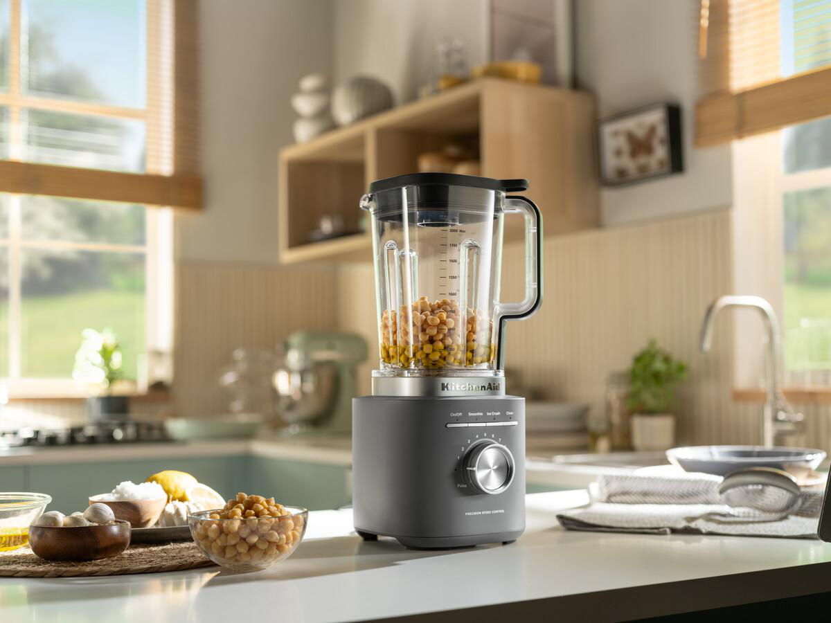 Ein KitchenAid-Blender mit Kichererbsen. Weitere Zutaten und Küchenutensilien befinden sich im Vorder- und Hintergrund.