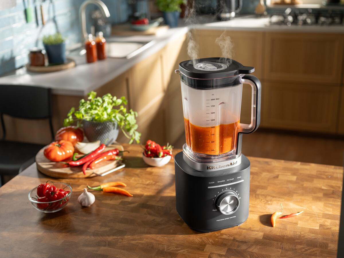 Ein Mixer mit orangener Flüssigkeit dampft auf einem Holztisch. Tomaten, Paprika und Knoblauch befinden sich in der Nähe. Küche im Hintergrund.