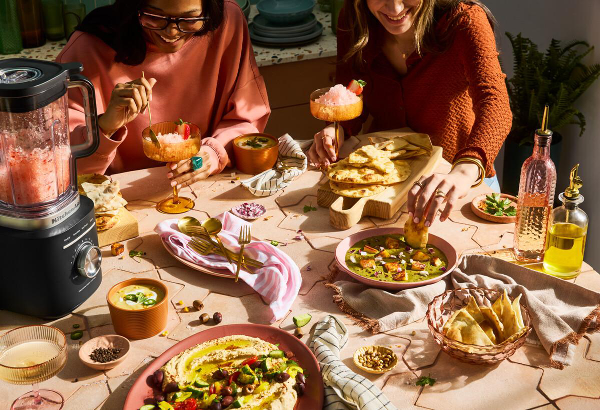 Zwei Frauen an einem Tisch mit Essen, Getränken und einem Mixer. Verschiedene Gerichte wie Hummus, Suppe und Naan sind verteilt.