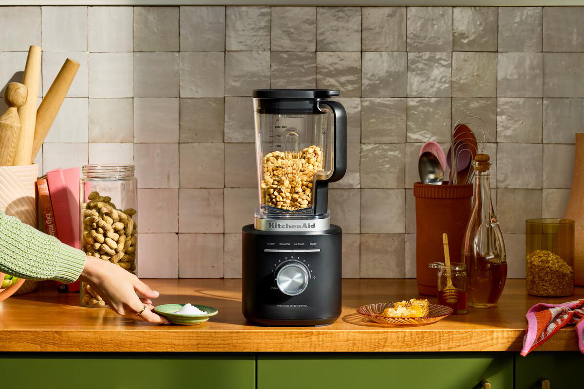 Ein KitchenAid-Mixer mit Erdnüssen steht auf einer Holztheke. Eine Hand greift nach einem Teller mit Salz. Honig und Brot sind ebenfalls vorhanden.