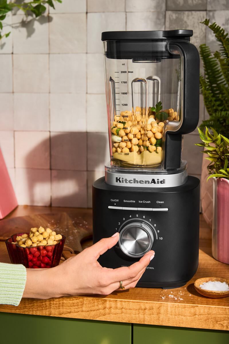 Ein schwarzer KitchenAid-Mixer wird mit Kichererbsen gefüllt. Eine Hand dreht den Drehknopf. Eine Schüssel mit Kichererbsen steht daneben.