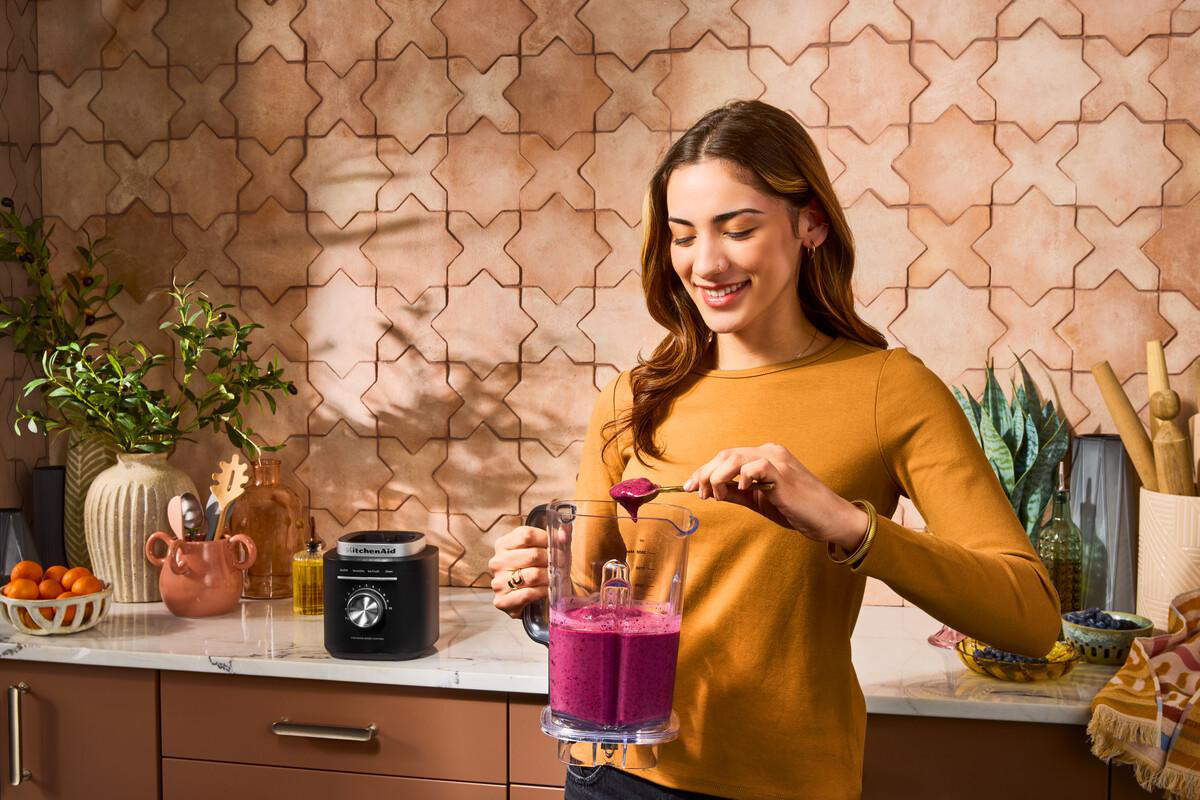 Frau bereitet Smoothie in der Küche zu. Sie hält einen Löffel über einem Mixer mit lila Smoothie.