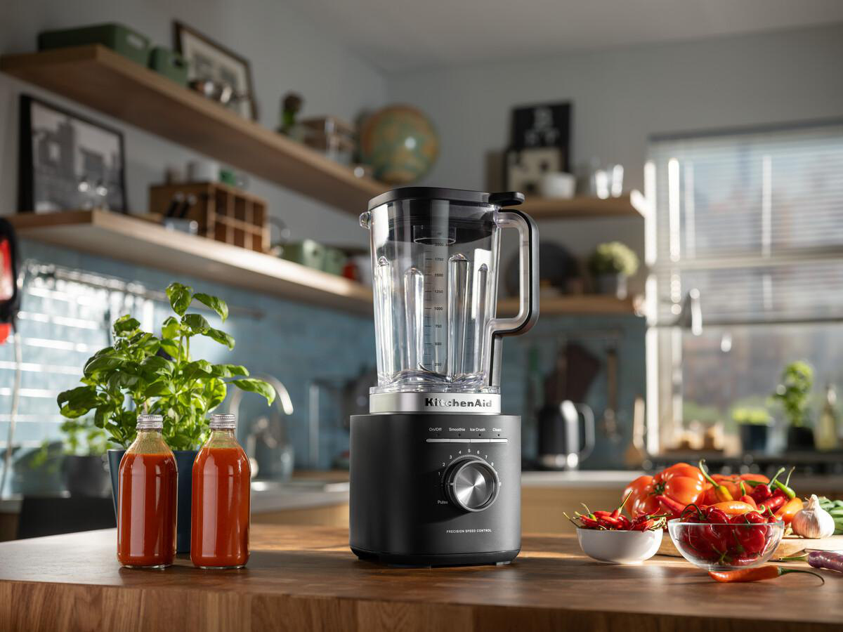 Ein schwarzer KitchenAid-Mixer steht auf einer Holztheke mit Gemüse. Flaschen mit Sauce und Basilikum befinden sich links. Tomaten und Paprika sind rechts.
