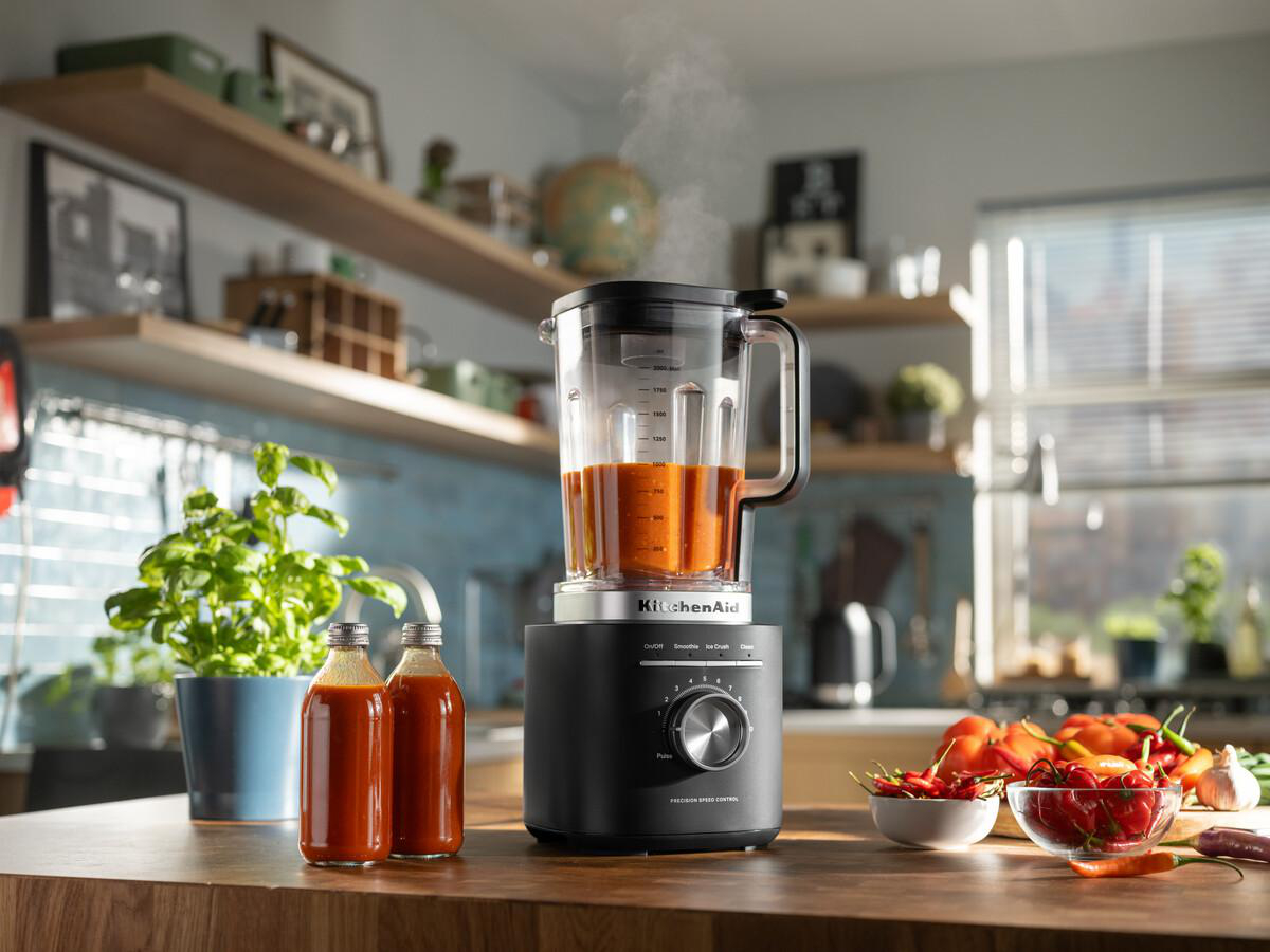 Eine Küchenszene mit einem schwarzen Mixer, der eine orangefarbene Flüssigkeit enthält. Flaschen mit Sauce, Pflanzen und Chilischoten stehen auf einer Holzoberfläche.